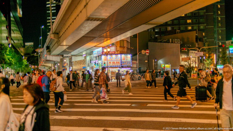 Pedestrians crossing a busy Osaka intersection beneath an elevated roadway at night.