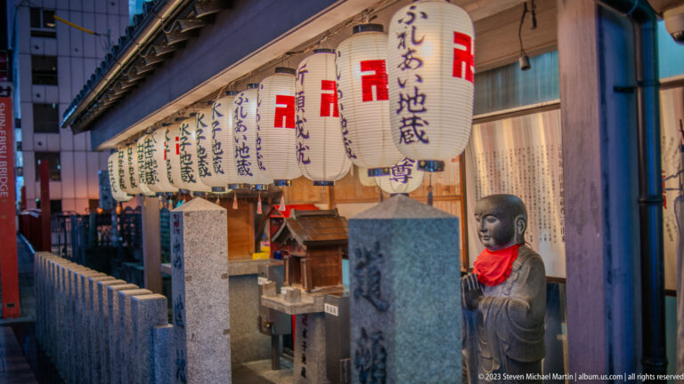 Stone Jizō statue with a red bib beside a lantern-lined roadside shrine in Osaka.