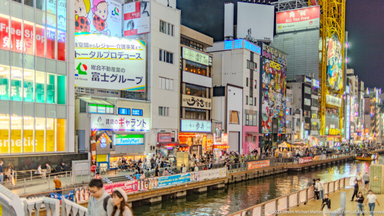 Dotonbori Canal with crowded promenades and brightly illuminated buildings at night.