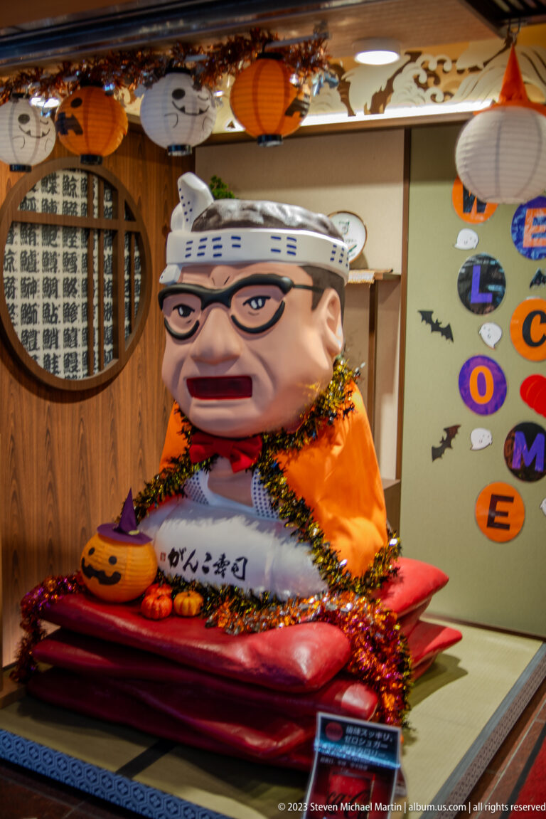 Large seated caricature figure in orange clothing and festive garland inside an Osaka venue.