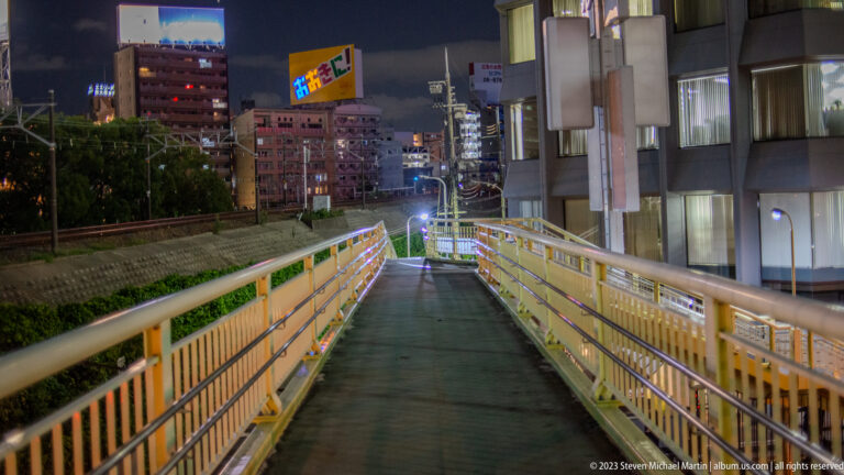 Yellow-railed pedestrian ramp descending toward illuminated buildings at night in Osaka.