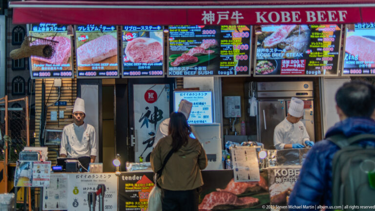 Cook serving customers at a Kobe beef counter beneath an illustrated menu board.