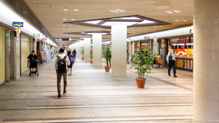 Bright underground Osaka shopping passage with pedestrians, columns, shops, and potted plants.