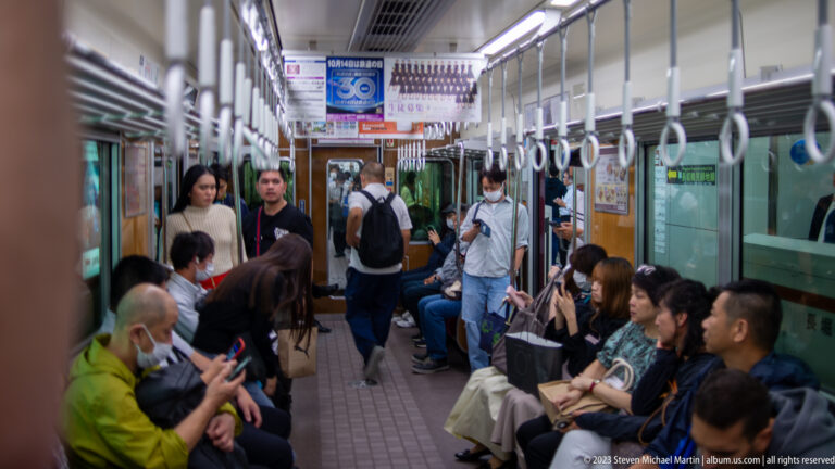 Passengers seated and standing inside an Osaka train beneath hanging straps.
