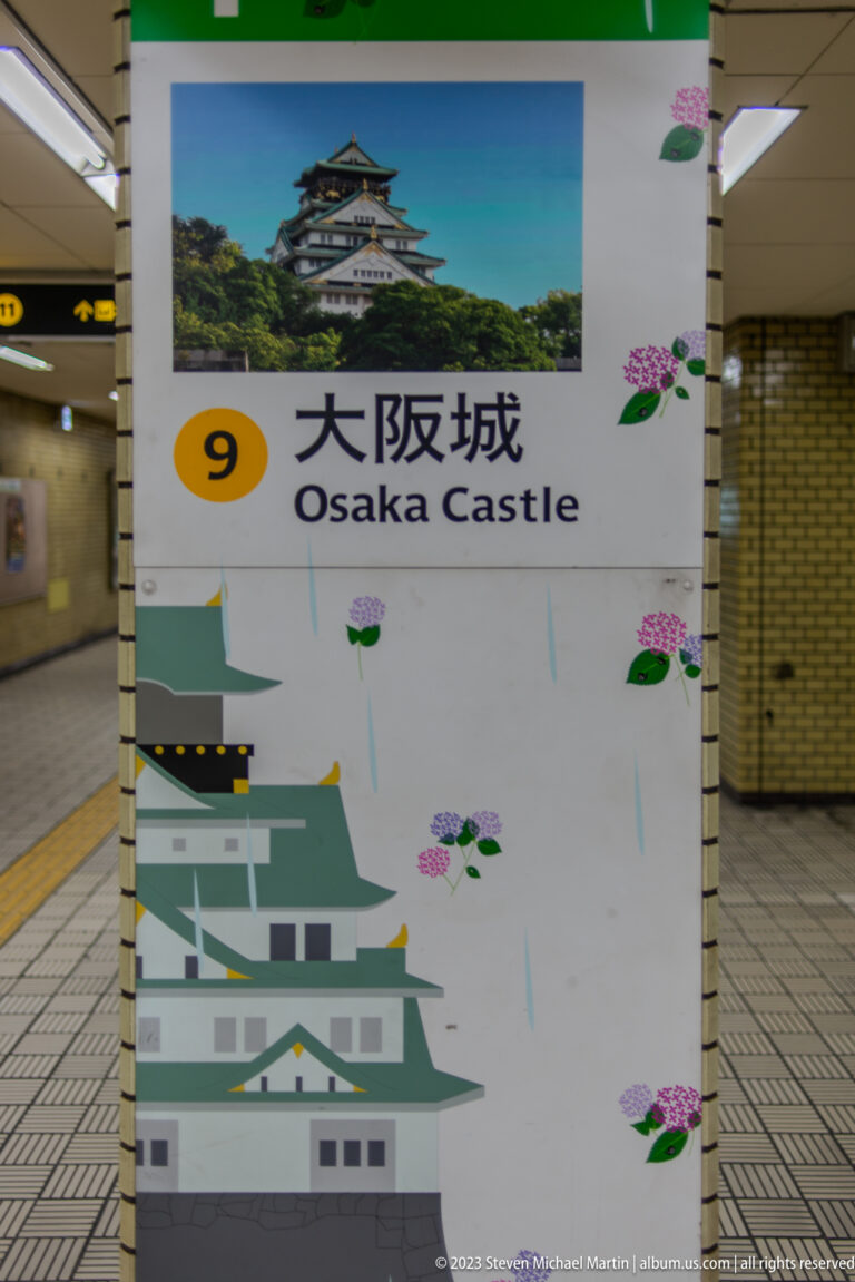 Station pillar sign reading Osaka Castle with a castle photograph and illustrated walls.