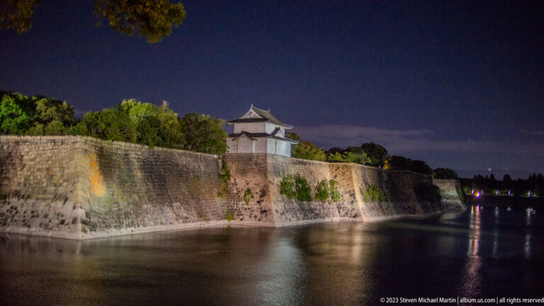 Illuminated Osaka Castle stone walls and white turret reflected in the moat at night.