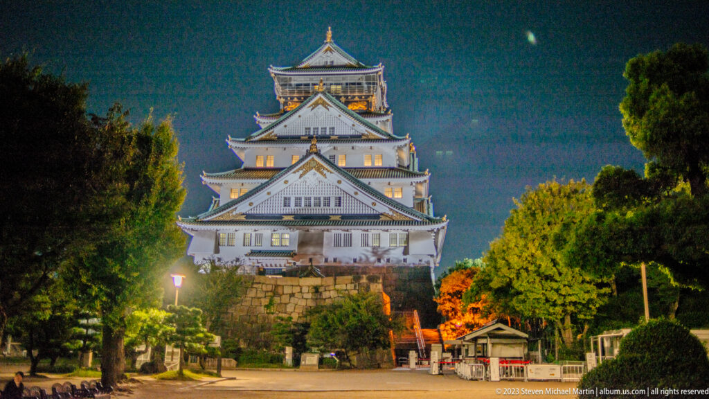 Illuminated Osaka Castle main keep viewed frontally across a tree-lined approach at night.
