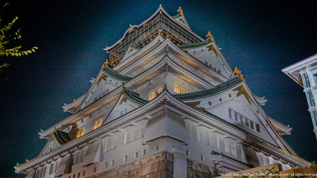 Low-angle corner view of Osaka Castle’s illuminated white walls and layered green roofs.