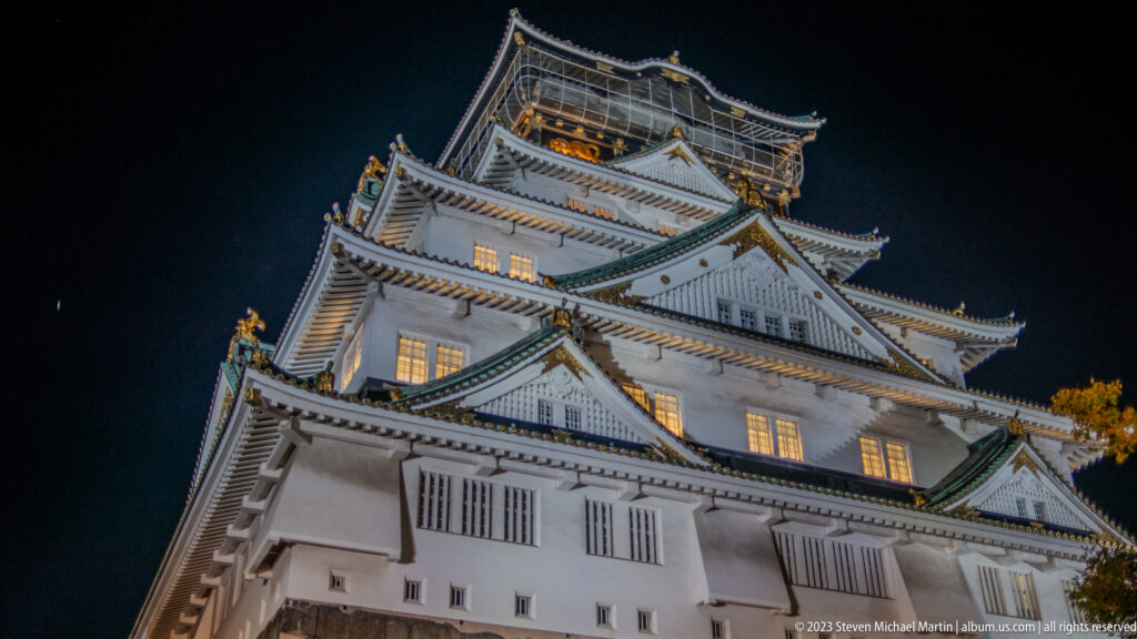 Oblique low-angle nighttime view of Osaka Castle’s tiered walls, roofs, and lit windows.