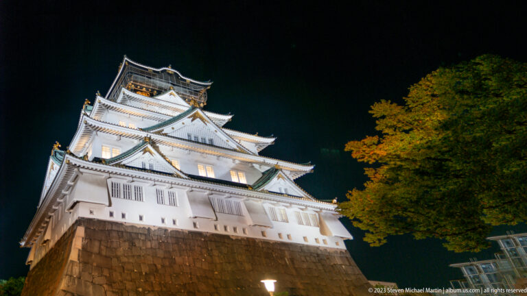 Osaka Castle illuminated above a stone foundation and a brightly lit tree at night.