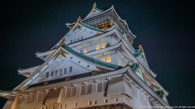 Close upward view of Osaka Castle gables, shutters, green eaves, and gilded roof ornaments.