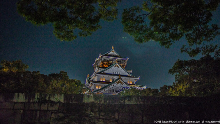 Distant illuminated Osaka Castle keep beyond dark stone walls and tree branches at night.