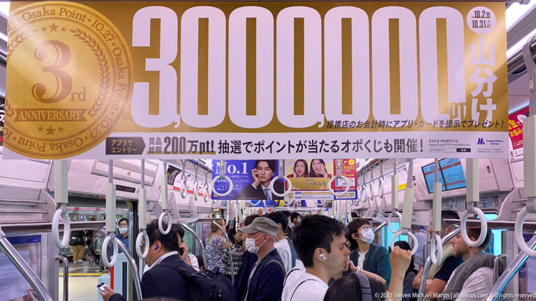 Crowded Osaka train car beneath a large advertisement reading three million yen.