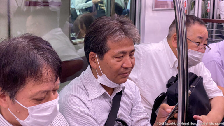 Three seated office workers, two masked, inside a crowded Osaka train.