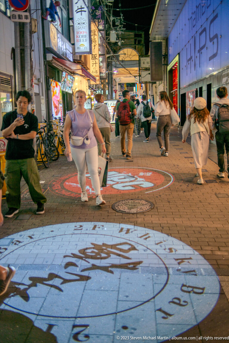 Pedestrians crossing a brightly lit Osaka nightlife street with painted pavement graphics.