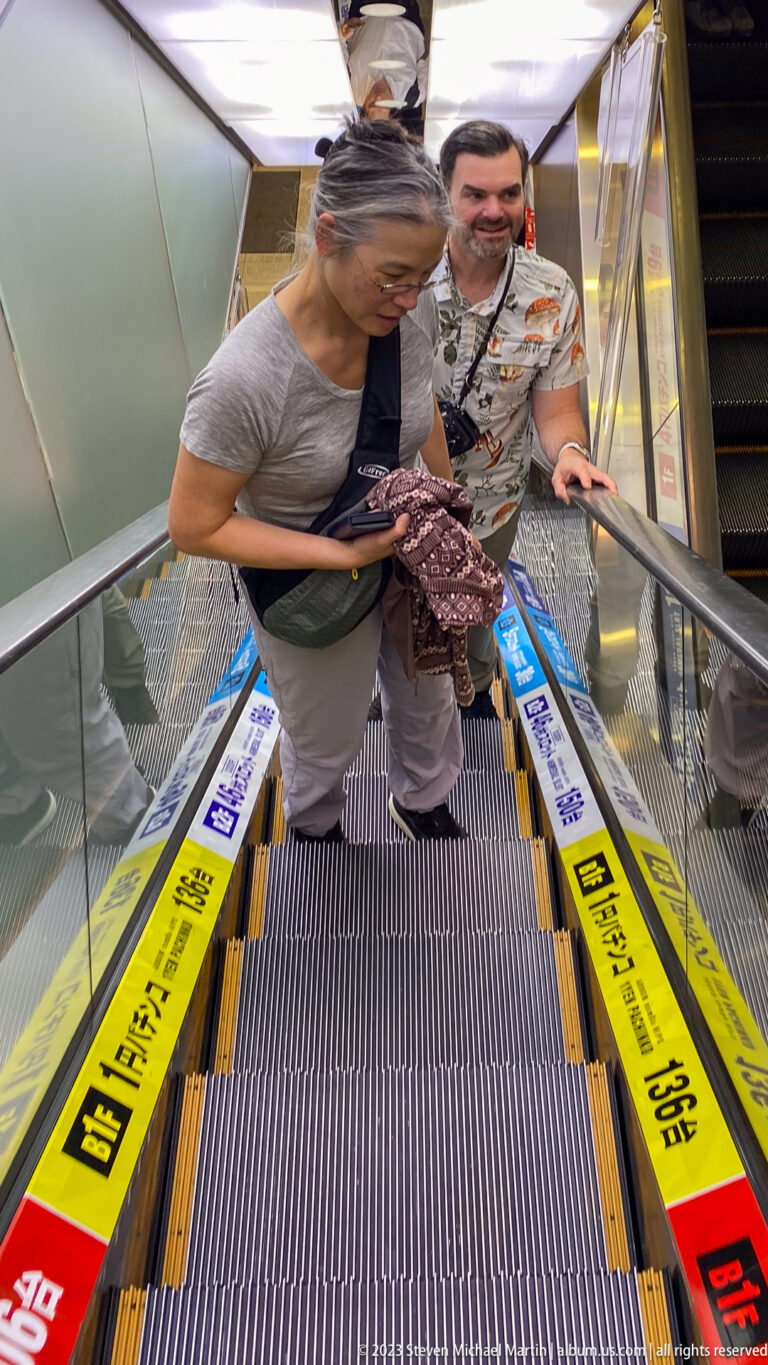 Two travelers ascending an indoor escalator lined with colorful Japanese advertising.