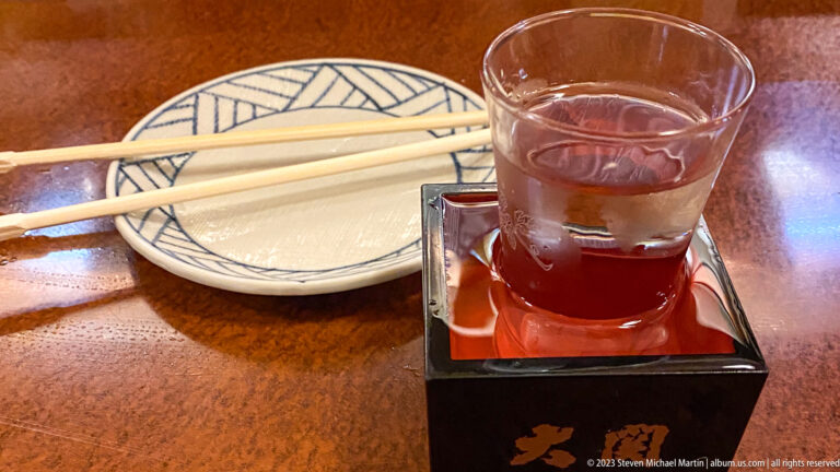 Glass of sake overflowing into a red-lacquered masu beside chopsticks and a plate.