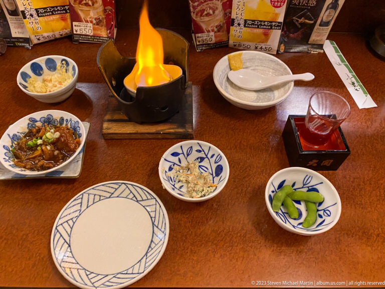 Yellow flame rising from a tabletop burner among plates, side dishes, and sake.
