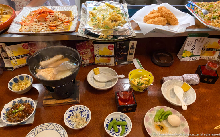 Overhead view of a Japanese counter meal with hot pot, fried food, vegetables, and sake.