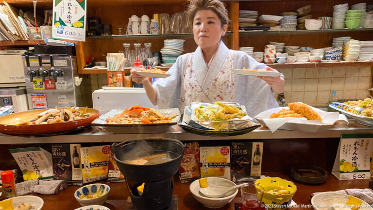 Server in a kimono-style uniform carrying plates behind a counter filled with dishes.