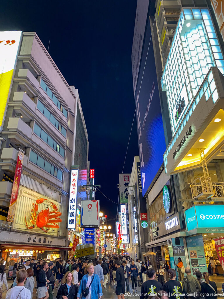Crowded Dotonbori street beneath the Kani Doraku crab sign and tall illuminated buildings.