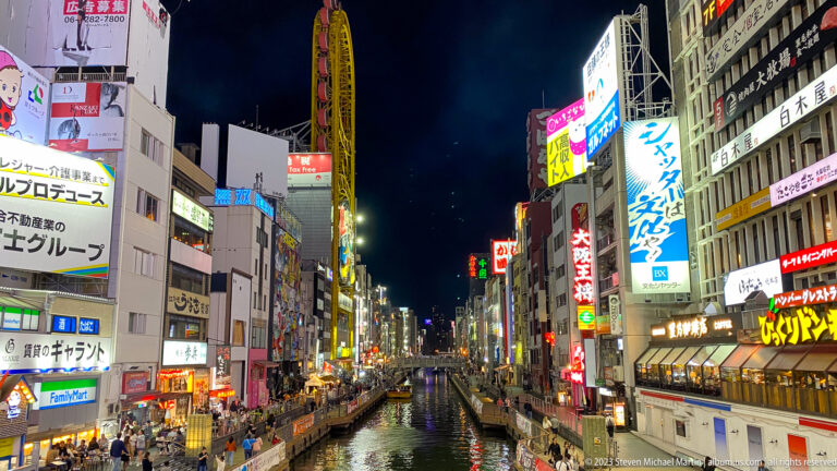 Long nighttime view along Dotonbori Canal between brightly lit commercial buildings.
