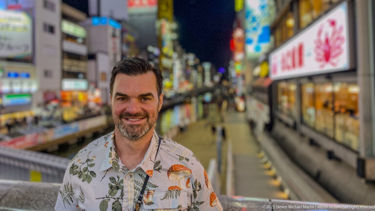 Smiling man in a patterned shirt posed above the blurred Dotonbori Canal promenade.