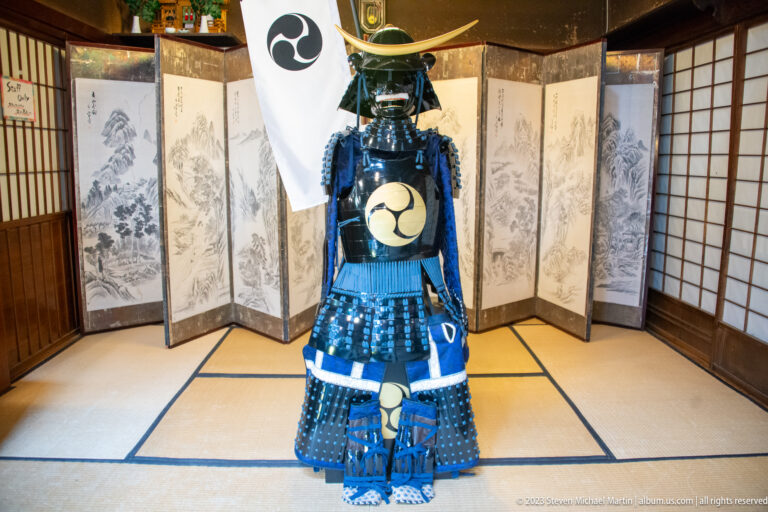 Blue-laced samurai armor stands on tatami mats before a painted folding screen and white heraldic banner.