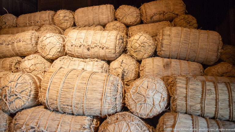 Stack of cylindrical rice bales wrapped in straw and bound with rope inside a dark storage room.