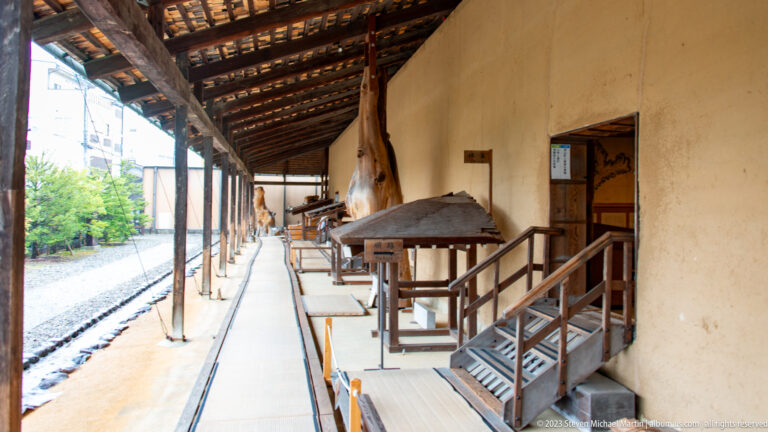 Long covered wooden passage beside a plaster wall with raised doors, steps, work platforms, and gravel yard.