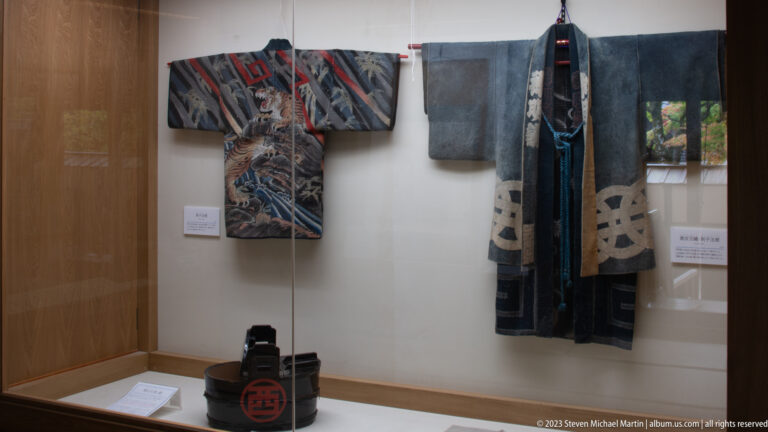 Two historic Japanese robes hang in a museum case above a black lacquered container with red crest.