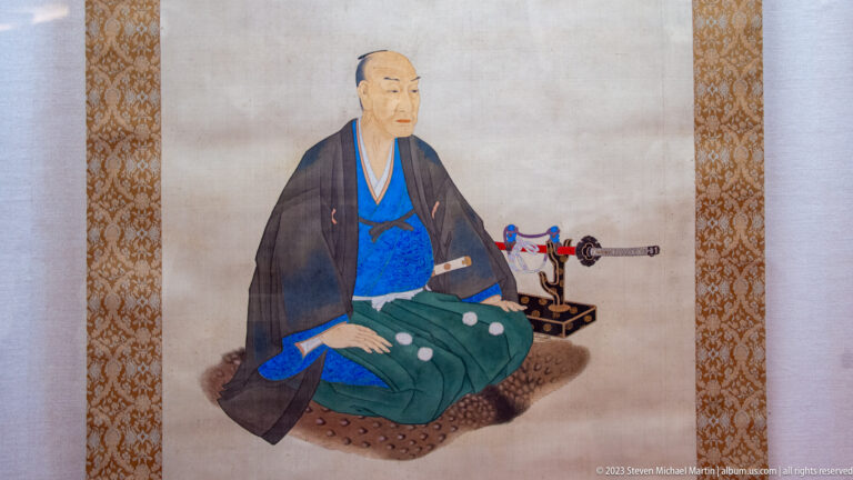 Painted seated Japanese official in formal robes beside a sheathed sword displayed on a stand.