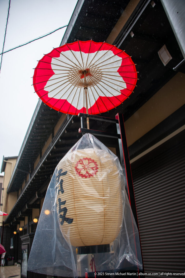 Red-and-white Japanese parasol hangs above a paper lantern wrapped in clear plastic beneath dark wooden eaves.