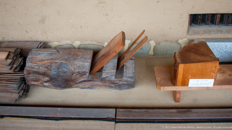 Traditional wooden planes, angled cutting tools, and stacks of thin roof shingles displayed on a work platform.