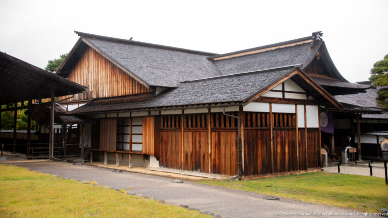 Large traditional timber hall with layered shingled roofs, white-plaster panels, exposed framing, and lawn in front.