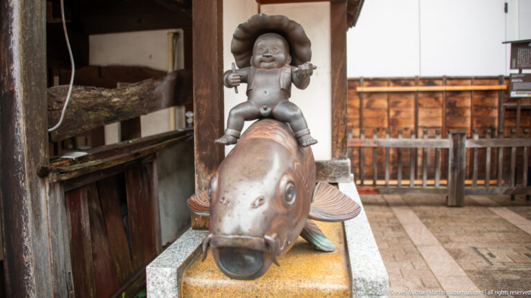 Small bronze childlike figure with large hair rides a stylized fish on a stone pedestal.