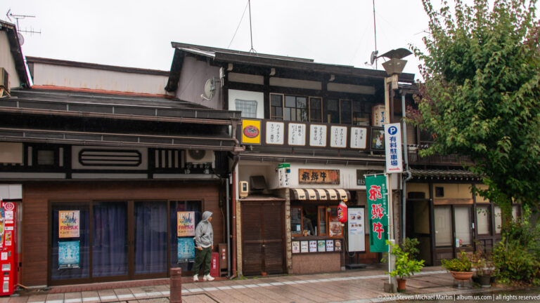 Traditional timber shopfronts with menu boards, banners, projecting signs, and layered rooflines at a Takayama street corner.