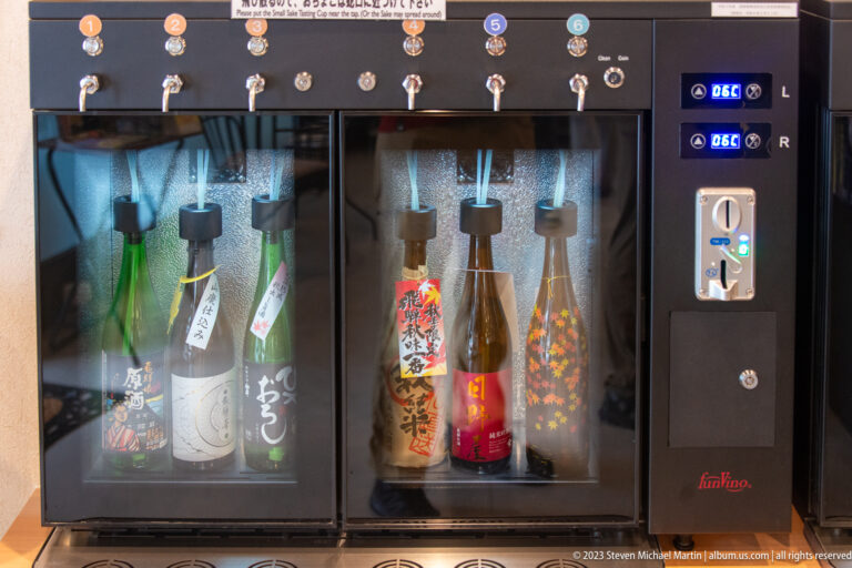 Refrigerated sake tasting machine with bottled selections behind glass, numbered taps, digital controls, and coin or card panel.
