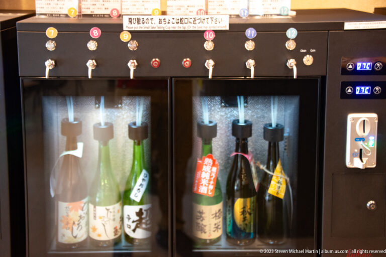 Five sake bottles stand in an automated refrigerated tasting dispenser beneath numbered taps and indicator lights.