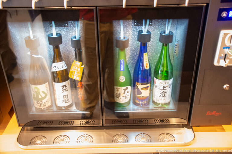 Six white, green, and blue sake bottles stand in two refrigerated tasting cabinets beneath dispensing heads.