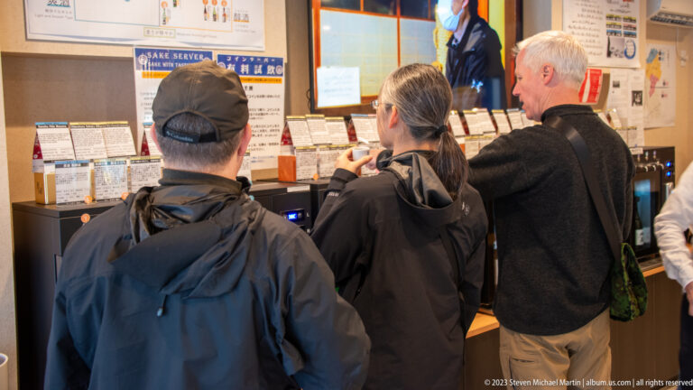 Visitors stand at a sake tasting counter reading selection cards and using automated wall-mounted dispensers.