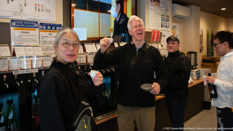 Two visitors hold small sake cups and react playfully in front of automated tasting dispensers and information cards.