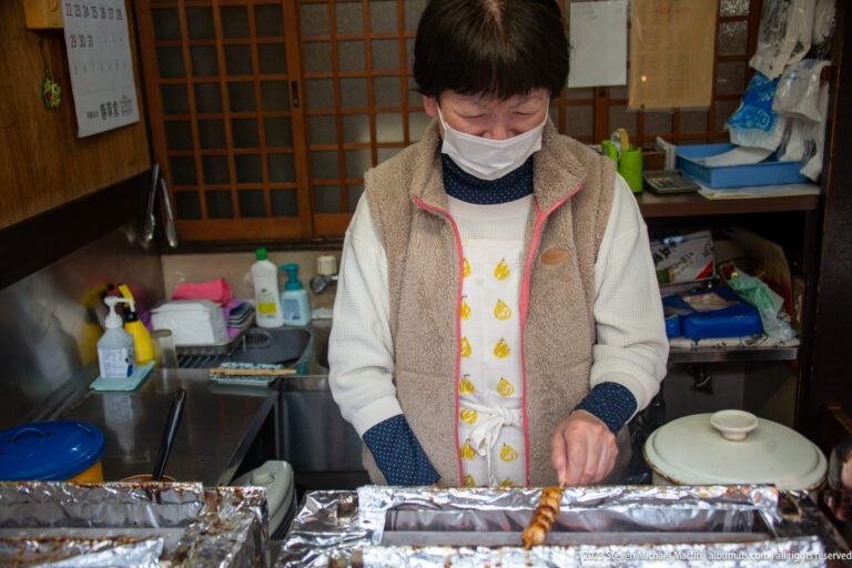 Masked shopkeeper grills a skewered rice snack over foil-lined burners inside a compact wooden food stall.