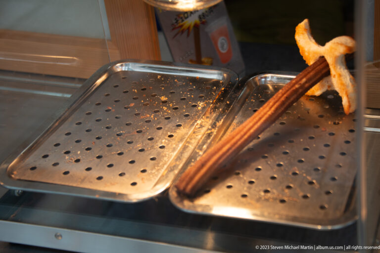 Freshly grilled irregular rice snack on a long wooden skewer held above perforated metal trays.