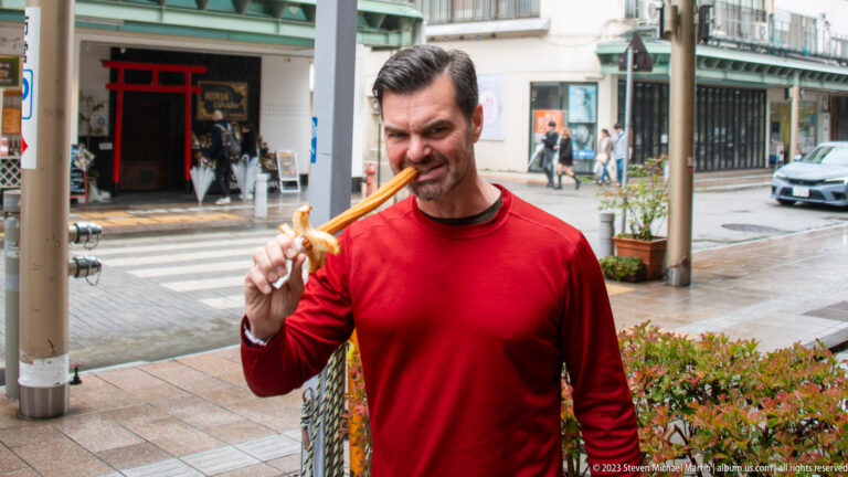 Man in red shirt raises a long skewer with grilled food toward his face on a wet Takayama street.