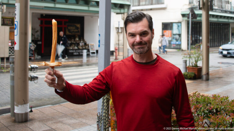 Man in red shirt holds a grilled snack on a long wooden skewer toward the camera on a wet street.