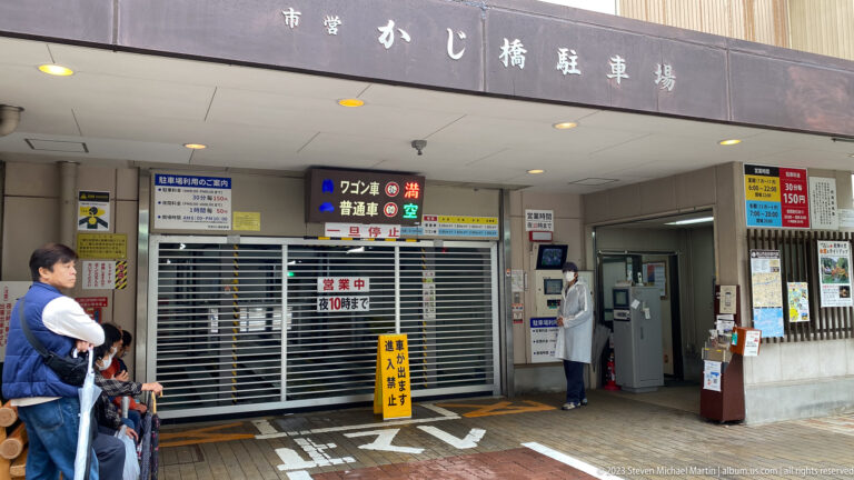 Entrance to a Japanese bicycle parking facility with signs, shuttered bays, and people waiting under a concrete canopy.