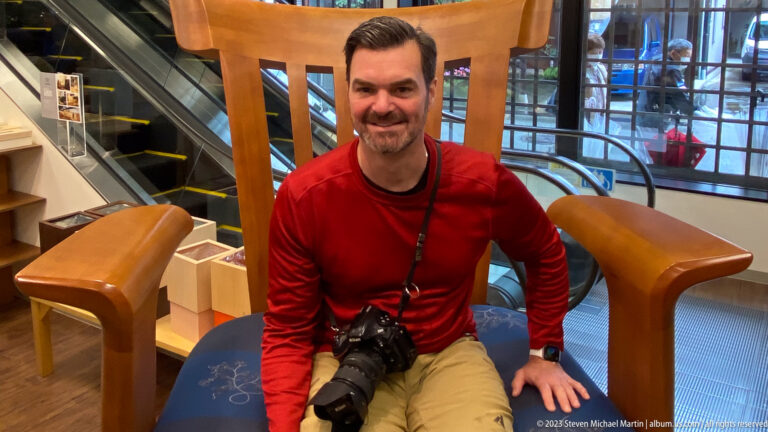 Smiling man in a red shirt seated indoors with a camera across his lap beside an escalator.
