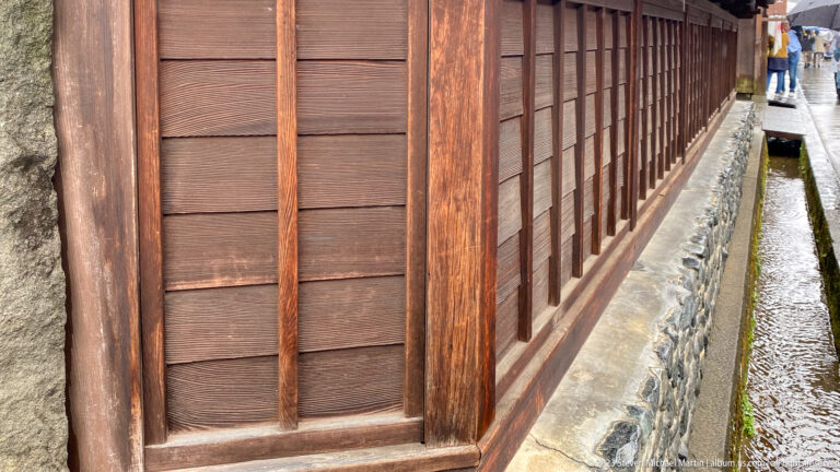 Close angled view of a weathered wooden lattice wall beside a narrow stone-lined water channel.