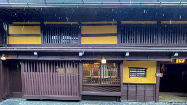 Dark wooden townhouse façade with lattice windows, deep eaves, and a warmly lit recessed shop entrance.